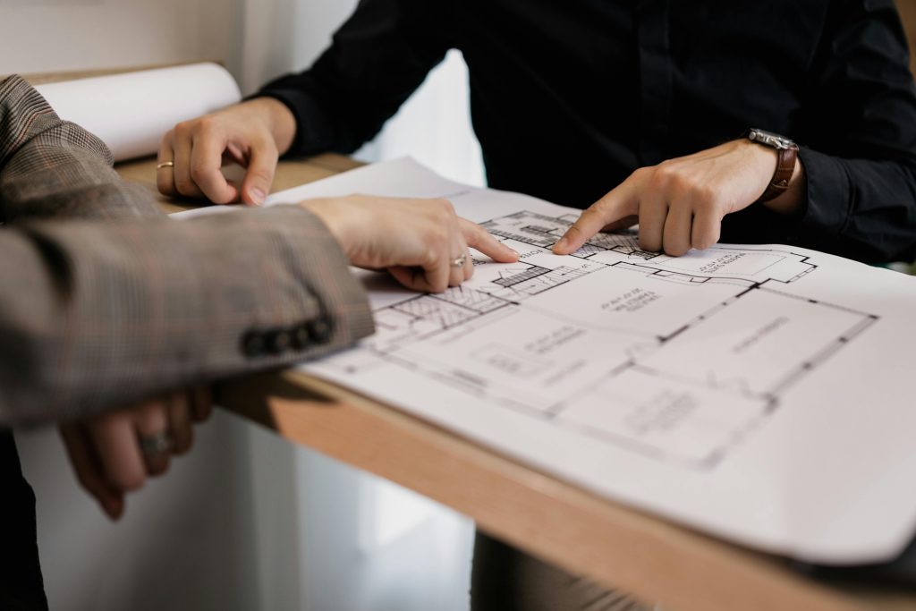 pexels photo 7642088 7642088 Two architects reviewing and pointing at a detailed floor plan on paper in an office setting.